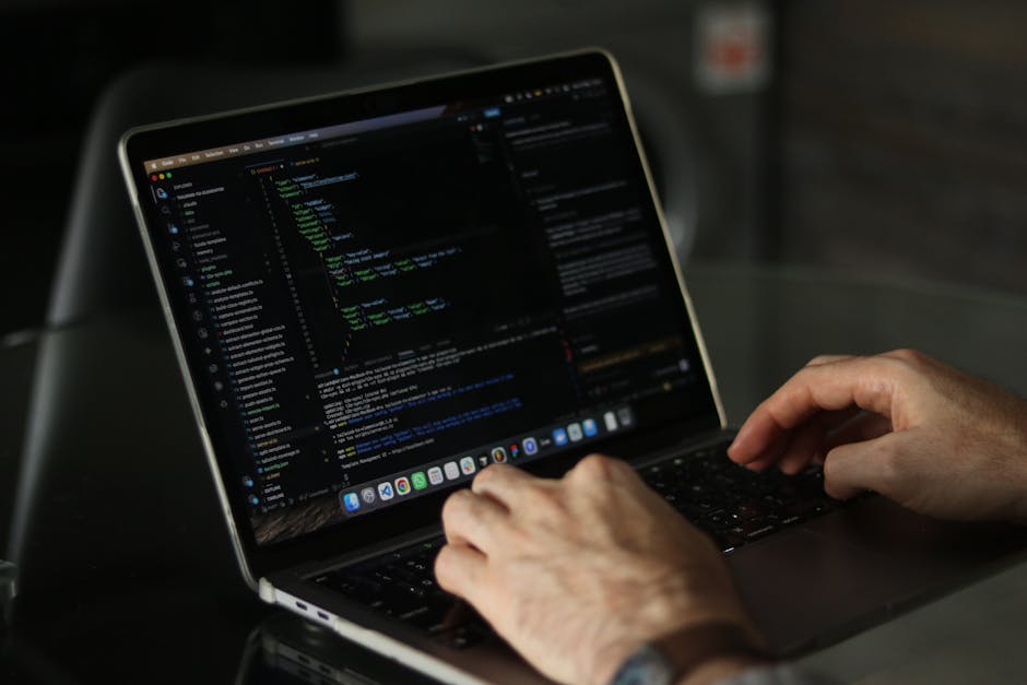 A pair of hands operating a sleek silver laptop with a dark screen displaying a code editor, suggesting a focus on programming or web development, with the background blurred to emphasize the device a